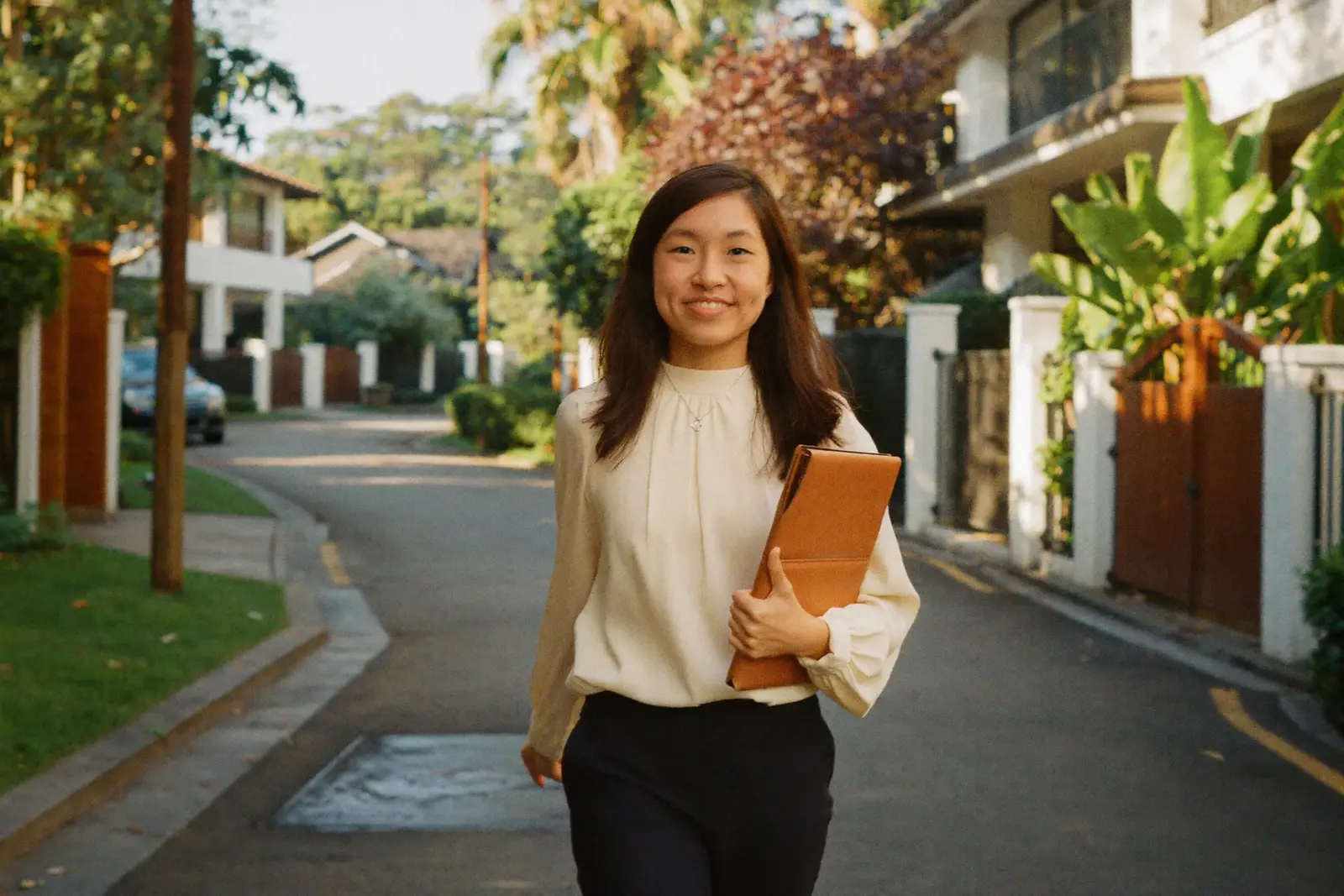 Jessin walking a leafy landed street in the golden hour, tan folio in hand.