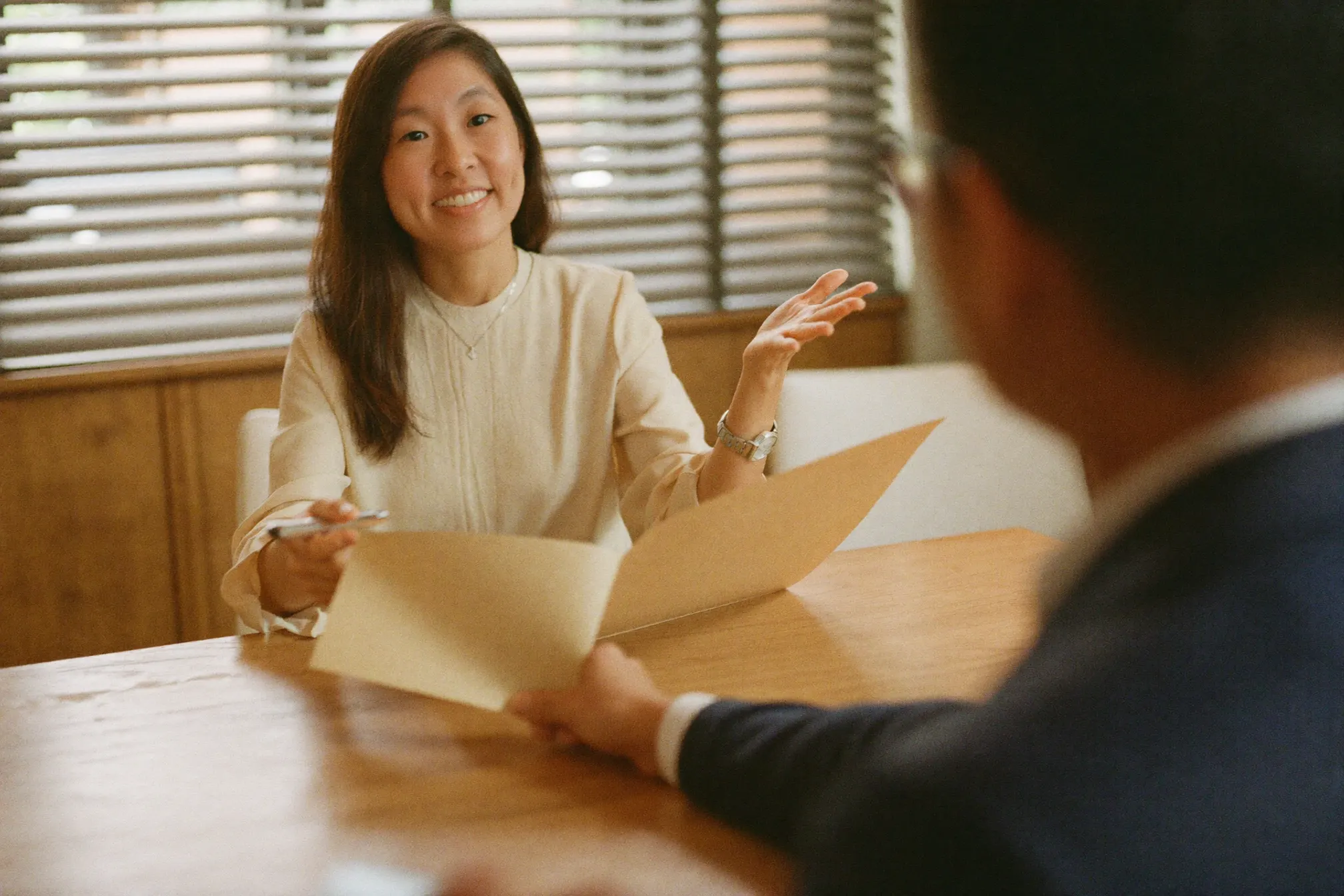 Jessin at a bright teak table, walking a client through an open kraft folder, pen in hand, louvred window light behind her. A representative scene.