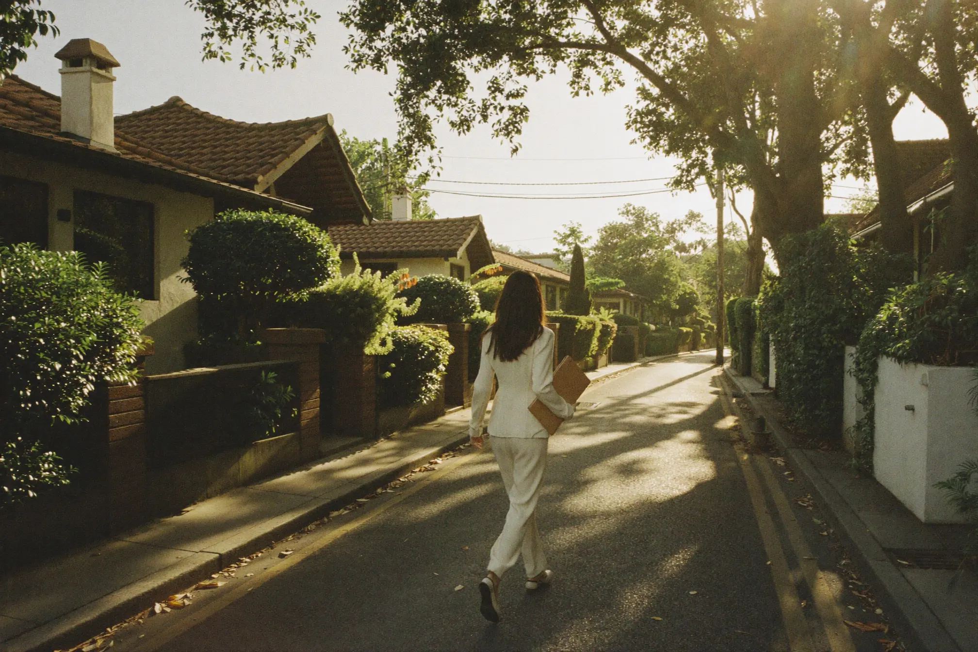 Back view of Jessin walking a leafy landed street in the golden hour, tan folio in hand.